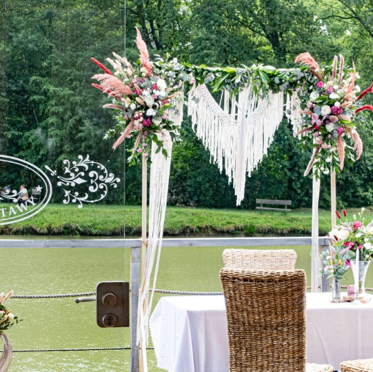 Udekorowane miejsce ceremonii ślubnej z kwiatami, makramą i widokiem na jezioro w tle.