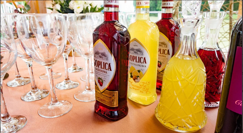 Ułożone na stole butelki likieru Soplica o smaku wiśniowym i pigwowym, obok karafka z żółtym trunkiem i kieliszki do wina.