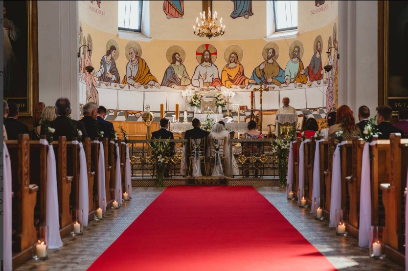 Para młoda klęczy przed ołtarzem w kościele, widok z perspektywy nawy głównej, czerwony dywan, ławki udekorowane białymi wstążkami i świecami.