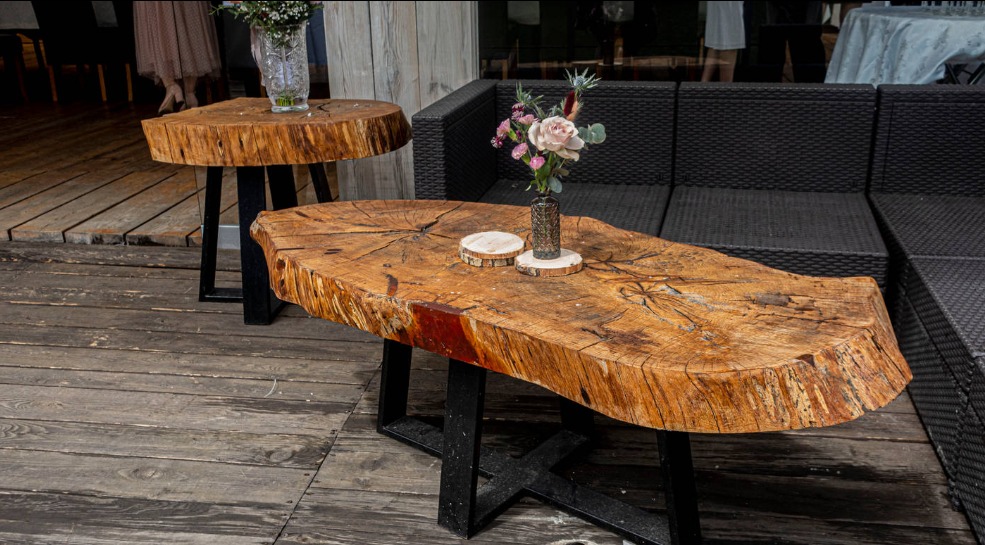 Dwa stoliki kawowe z plastrów drewna na czarnych, metalowych nogach, jeden z bukietem kwiatów, na drewnianym tarasie obok czarnej sofy.