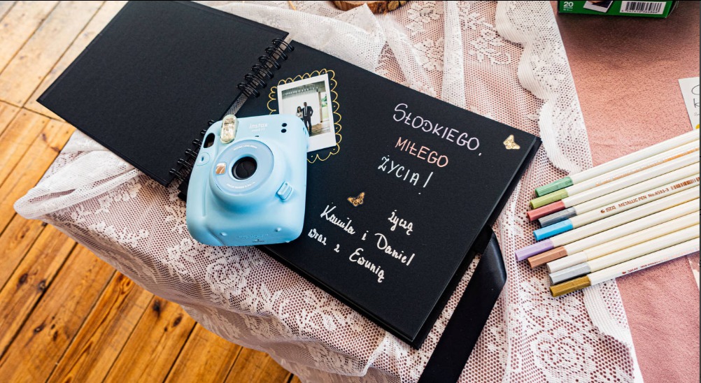 Niebieski aparat Instax leżący na otwartym, czarnym albumie z życzeniami ślubnymi napisanymi srebrnym pisakiem, obok kolorowe kredki i zdjęcie pary młodej w ramce.