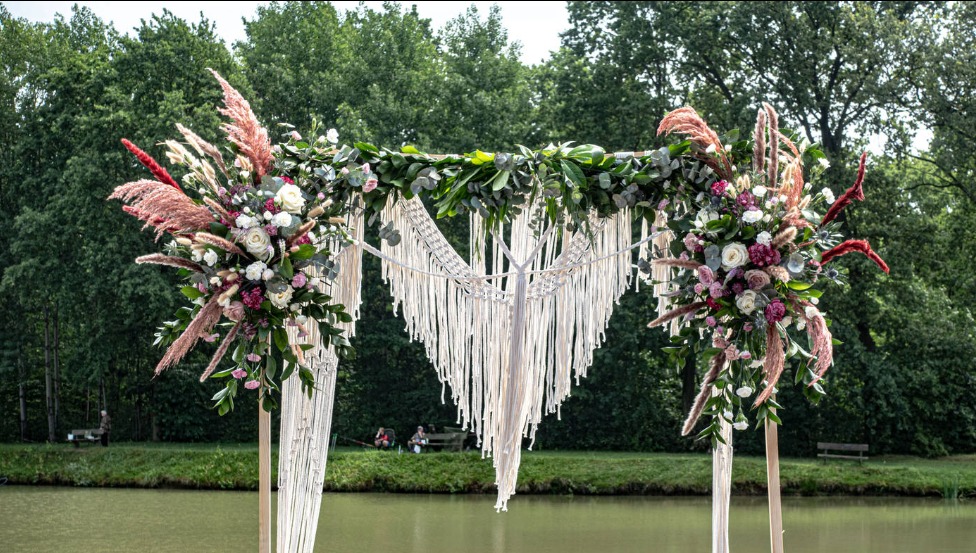 Dekoracja ślubna w stylu boho z makramą i kwiatami, ustawiona nad brzegiem stawu, w tle widoczni ludzie na ławce.
