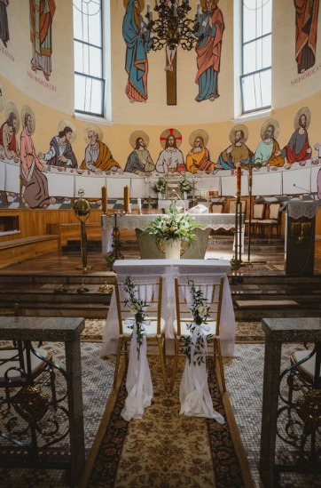 Wnętrze kościoła przygotowane na uroczystość ślubną, z dekoracjami kwiatowymi na krzesłach i ołtarzu, freski z motywami religijnymi na ścianach.
