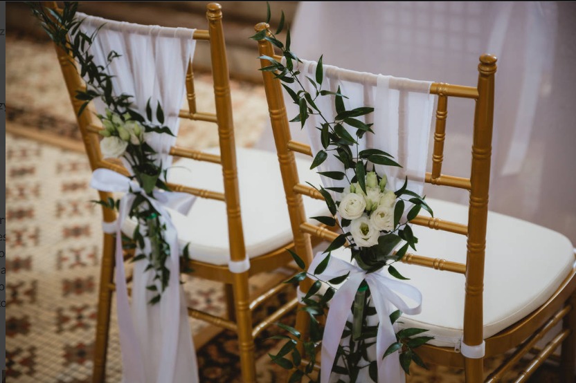 Złote krzesła udekorowane białymi kwiatami, zielonymi gałązkami i zwiewnymi białymi wstążkami, przygotowane na uroczystość.