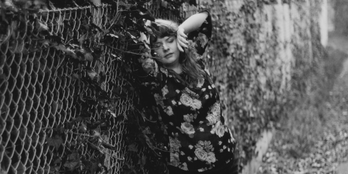 Portret kobiety w kwiecistej bluzce, opierającej się o zarośnięte ogrodzenie z siatki w czarno-białej tonacji.