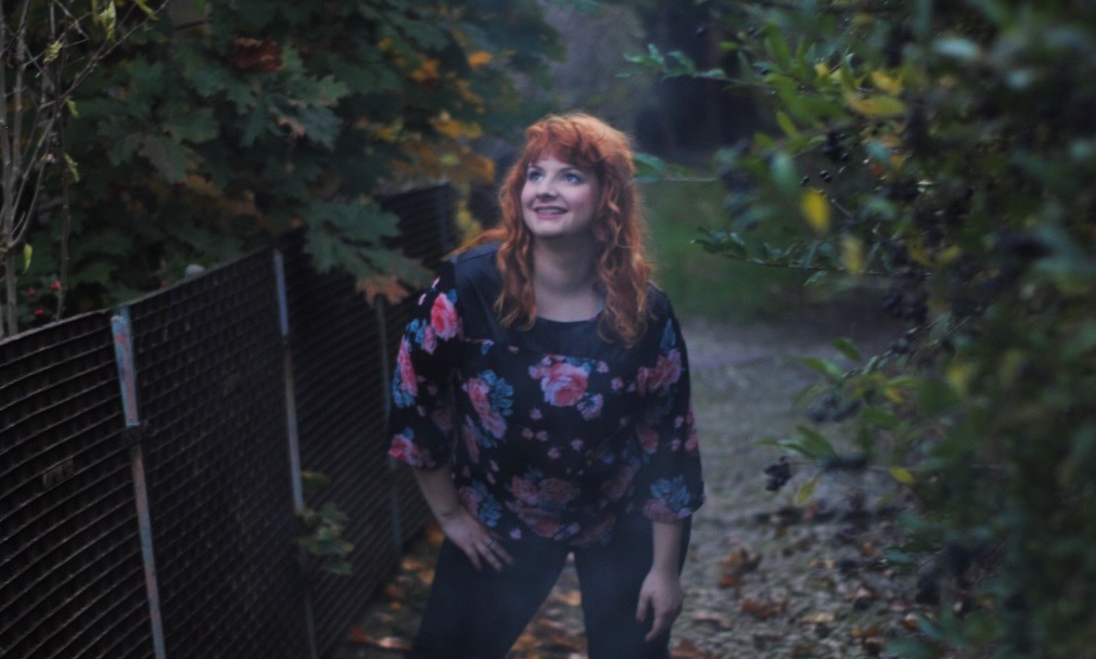 Kobieta z rudymi włosami w bluzce w kwiaty, stojąca na ścieżce w otoczeniu zieleni, spoglądająca w górę w lewo.