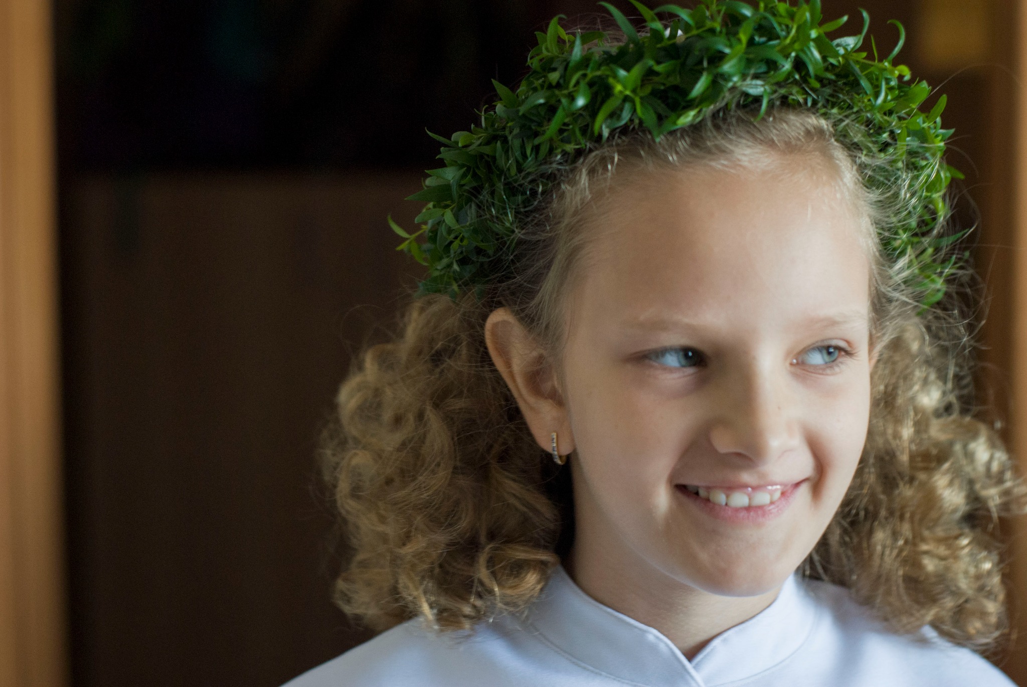 Portret uśmiechniętej dziewczynki z wiankiem z zielonych liści na głowie, skierowanej wzrokiem w lewo, z widoczną złotą kolczykiem w uchu.