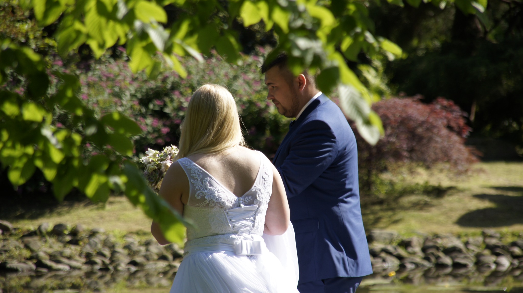 Para młoda w plenerze: panna młoda w białej sukni z bukietem, pan młody w granatowym garniturze, zielone tło z drzewami i krzewami, ujęcie zza liści.