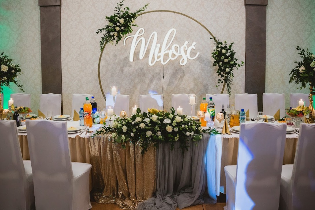 Uroczysty stół weselny z dekoracją kwiatową, świecami, złotym obrusem z cekinami i napisem 'Miłość' w okręgu.