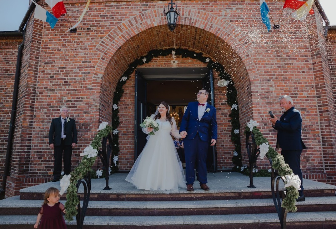 Para młoda wychodzi z kościoła pod deszczem konfetti, na schodach stoi mała dziewczynka w bordowej sukience, w tle ceglana fasada budynku z dekoracją kwiatową i girlandami.