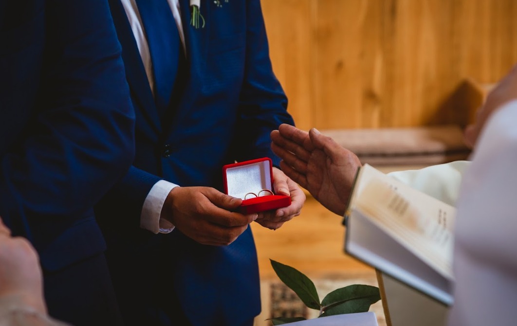 Wymiana obrączek podczas ceremonii ślubnej w kościele, zbliżenie na dłonie trzymające czerwone pudełko z obrączkami, widoczny fragment księdza.