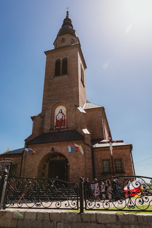 Kościół z czerwonej cegły z wieżą i obrazem Jezusa, udekorowany flagami, przed wejściem zgromadzeni ludzie.