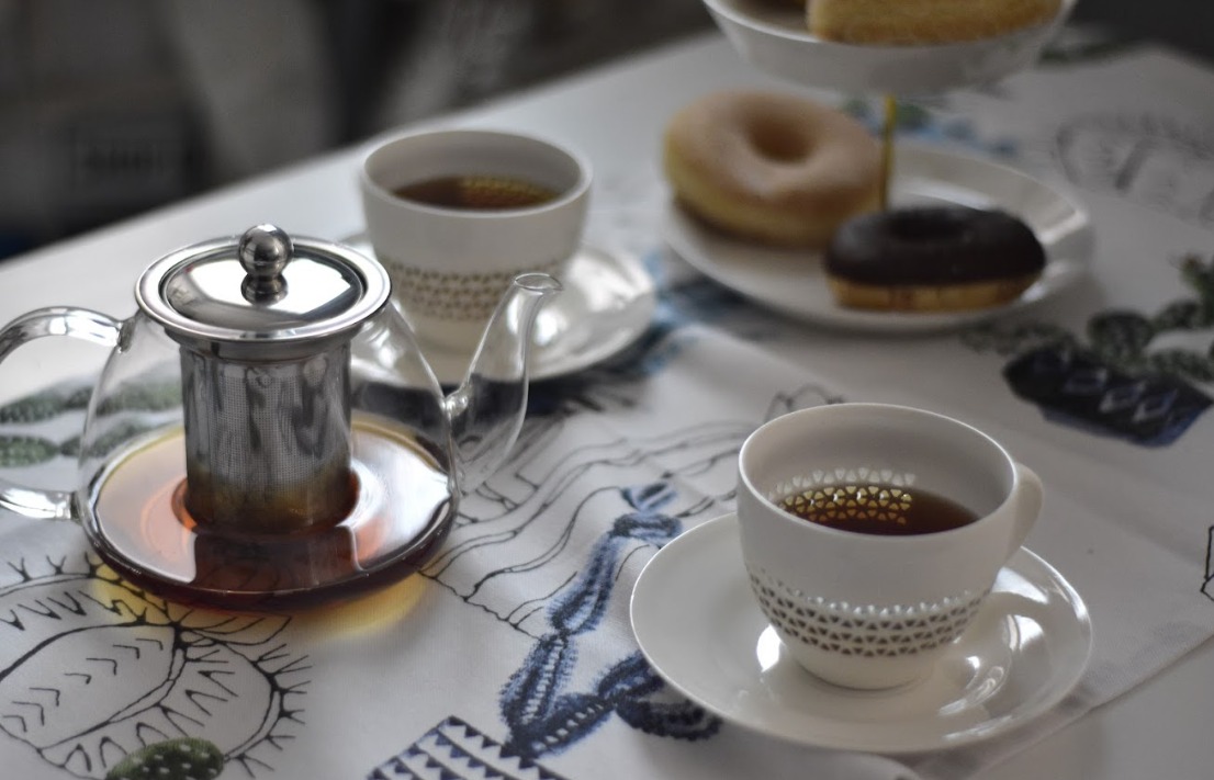 Szklany czajnik z zaparzaczem, filiżanki z herbatą i talerz z pączkami na wzorzystym obrusie w neutralnej tonacji.