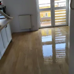 Błyszcząca, świeżo polakierowana podłoga z drewna, odbijająca światło okien balkonowych, z widocznym fragmentem białej zabudowy kuchennej w tle.