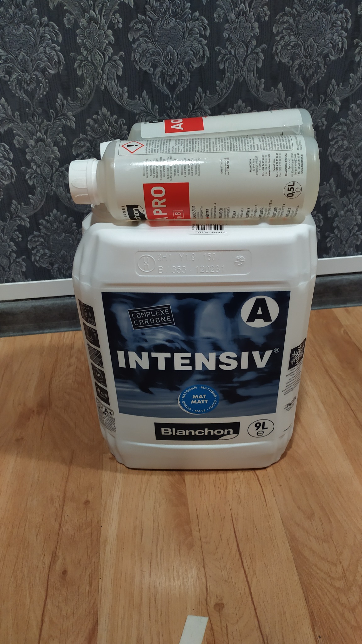 Kanister 9L z intensywnym środkiem do parkietów Blanchon Intensiv VL Mat, na nim dwie butelki 0.5L Blanchon Aquapro B, na drewnianej podłodze, w tle wzorzysta tapeta.