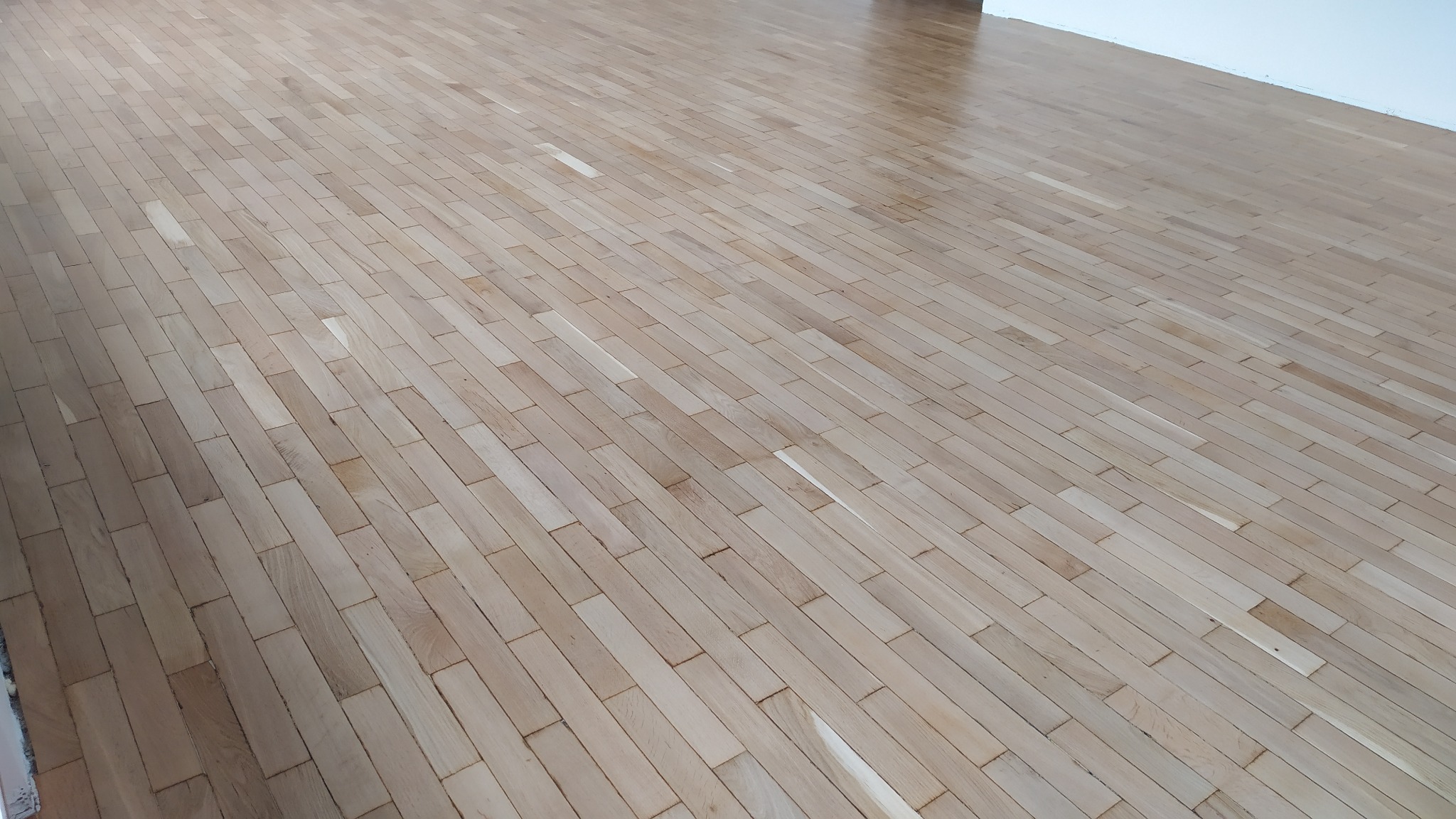 Jasny, lakierowany parkiet ułożony w jodełkę, widoczny z perspektywy pod lekkim kątem, z minimalnym odblaskiem światła w górnej części kadru.