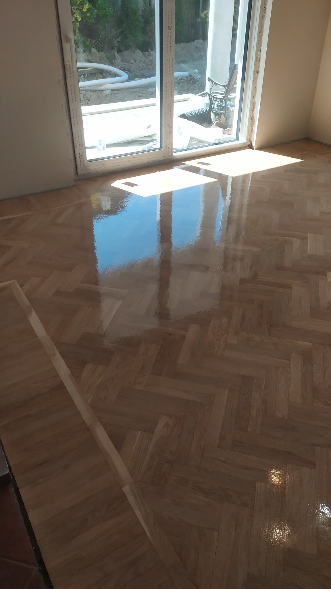 Świeżo ułożony, lakierowany parkiet w jodełkę, z widocznym odbiciem okna i fragmentem listwy przypodłogowej w rogu pomieszczenia.