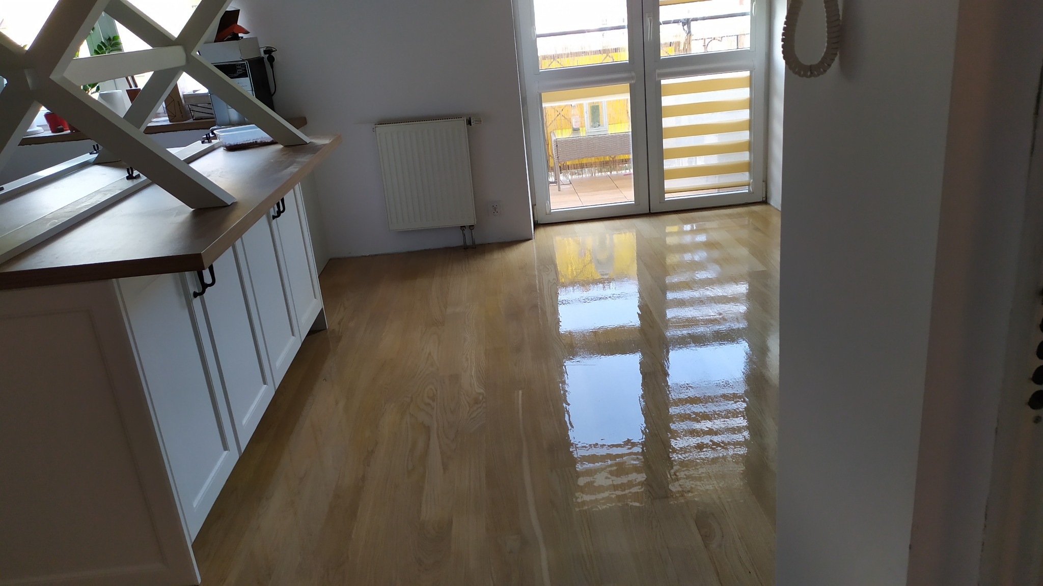 Błyszcząca, świeżo polakierowana podłoga z drewna, odbijająca światło okien balkonowych, z widocznym fragmentem białej zabudowy kuchennej w tle.