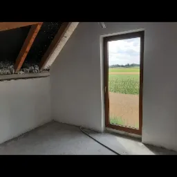 Wnętrze poddasza w trakcie prac wykończeniowych, widoczne surowe ściany, drewniana konstrukcja dachu z izolacją, okno z widokiem na pole i wężyk na podłodze.