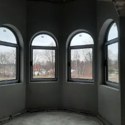 Wnętrze w stanie deweloperskim z trzema łukowymi oknami z widokiem na zabudowę jednorodzinną w pochmurny dzień, ściany pokryte świeżym tynkiem.