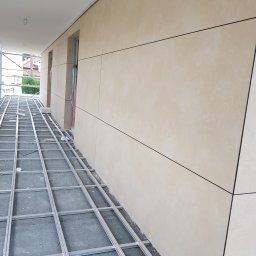 Jakub Polcyn - Montaż elewacji wentylowanej z płyt w kolorze beżowym, widoczne metalowe profile mocujące i fragment rusztowania.