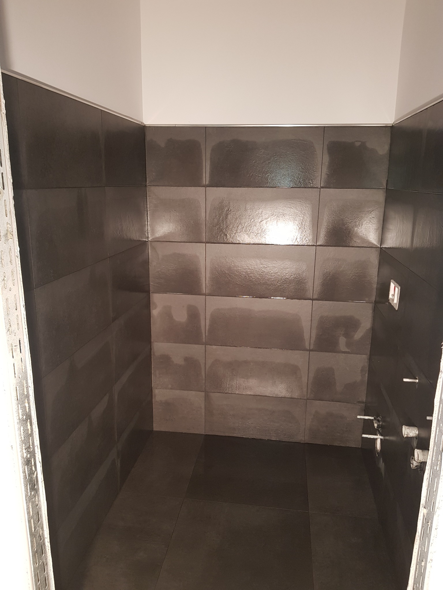 Świeżo ułożone, ciemnoszare płytki w małej łazience z widocznymi przyłączami hydraulicznymi i listwą wykończeniową przy suficie.