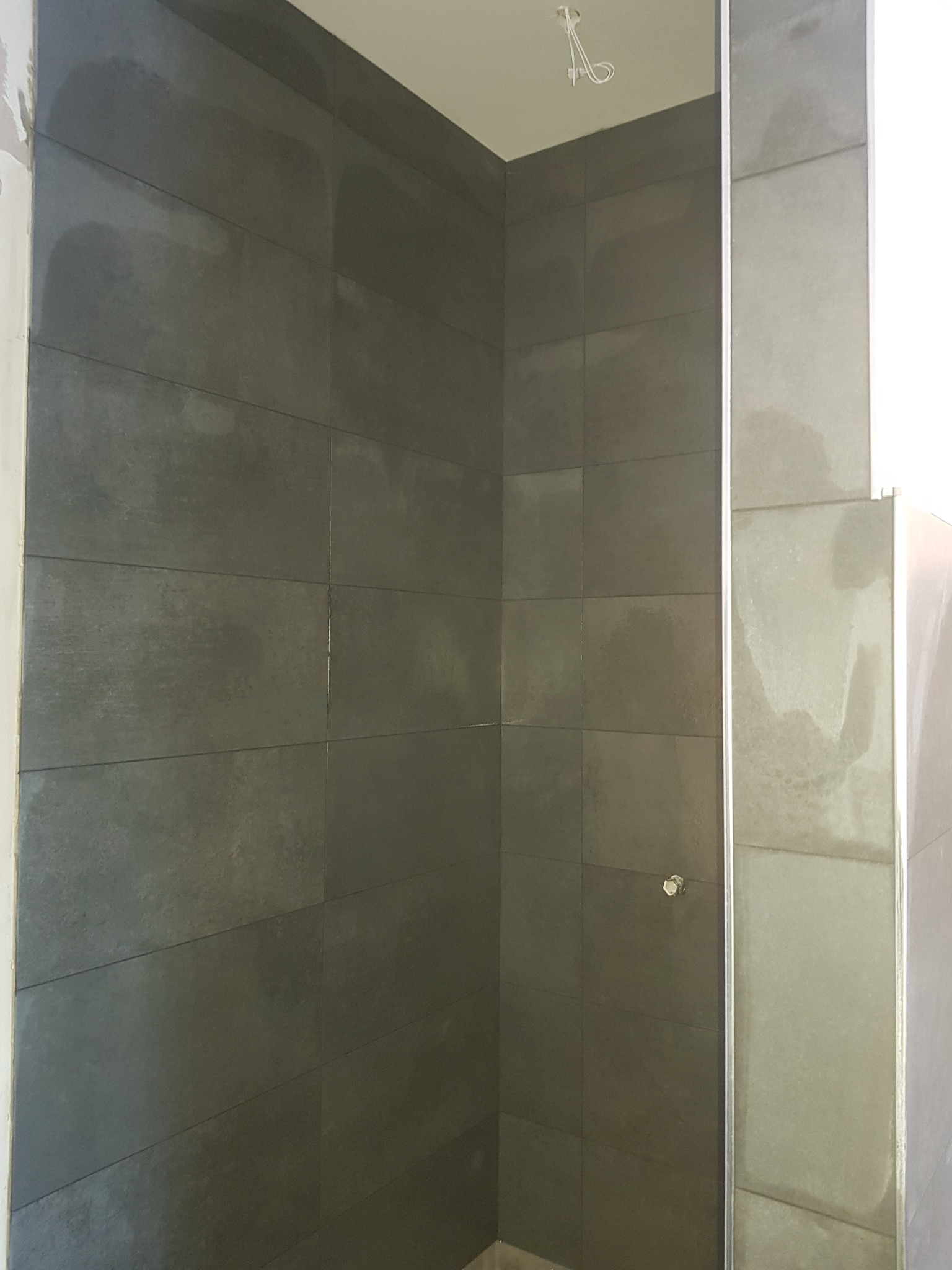 Ciemnoszare, matowe płytki w kabinie prysznicowej, widoczny fragment szklanej ściany prysznica i wystające kable oświetleniowe.