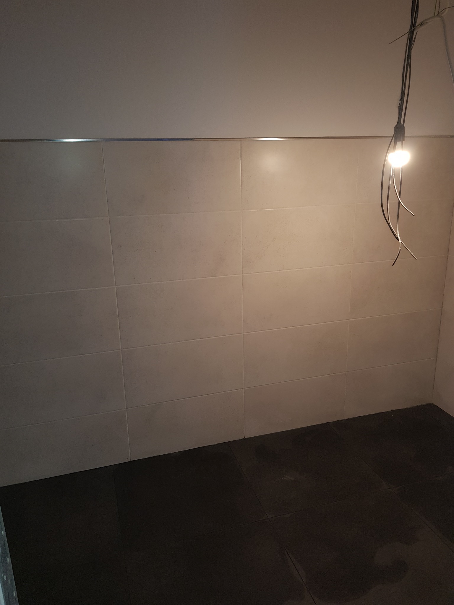 Ściana wyłożona jasnymi, kwadratowymi płytkami z metalową listwą wykończeniową pod sufitem oraz ciemna podłoga z płytek, oświetlona wiszącą żarówką na kablach.