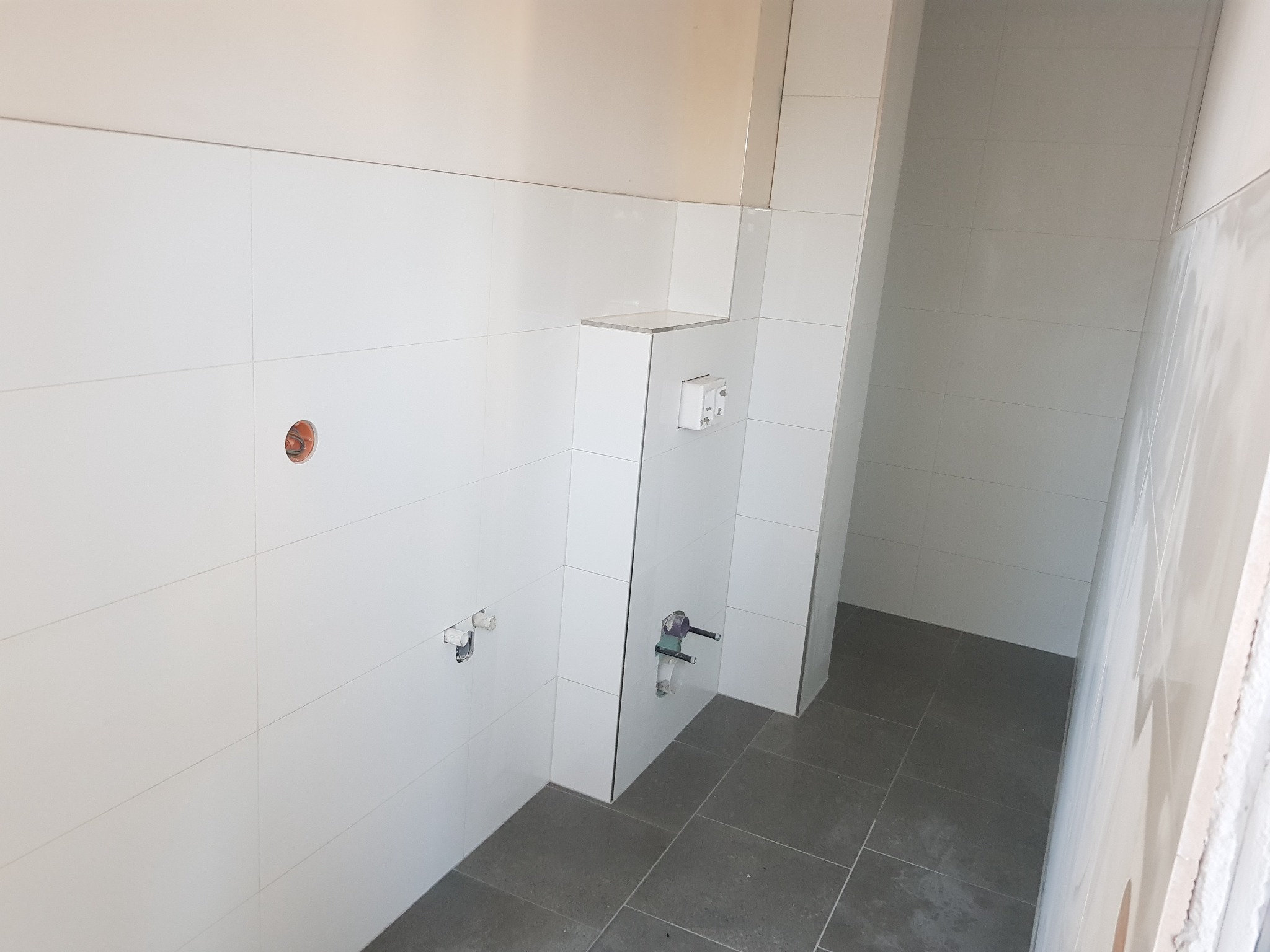 Białe, kwadratowe płytki ścienne i szare płytki podłogowe w trakcie montażu w łazience, widoczne podejścia wodne i gniazdko elektryczne.