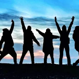Sylwetki grupy osób świętujących na plaży o zachodzie słońca, jedna osoba wykonuje wyskok, pozostali z podniesionymi rękami.