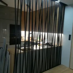 Nowoczesna kuchnia z drewnianymi blatami, oddzielona od reszty pomieszczenia czarną, pionową, ażurową konstrukcją. Widoczne płyty do montażu oparte o ścianę.