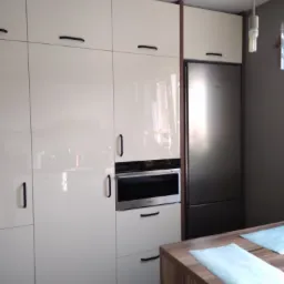 Biała, lakierowana zabudowa kuchenna z czarnymi, minimalistycznymi uchwytami, wbudowany piekarnik i lodówka w stali nierdzewnej, wyspa kuchenna z blatem w kolorze drewna.