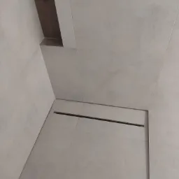 Szare płytki w kabinie prysznicowej z odpływem liniowym, widok z góry pod kątem, z fragmentem drewnianego elementu dekoracyjnego na suficie.