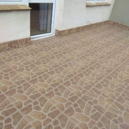 Remonty Lucas - Balkon wyłożony brązowymi płytkami imitującymi kamień o nieregularnym kształcie, z widocznymi białymi drzwiami balkonowymi i oknem po prawej stronie.