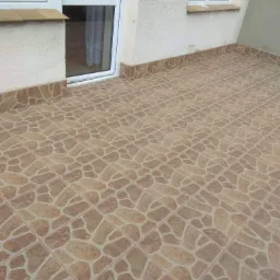 Balkon wyłożony brązowymi płytkami imitującymi kamień o nieregularnym kształcie, z widocznymi białymi drzwiami balkonowymi i oknem po prawej stronie.