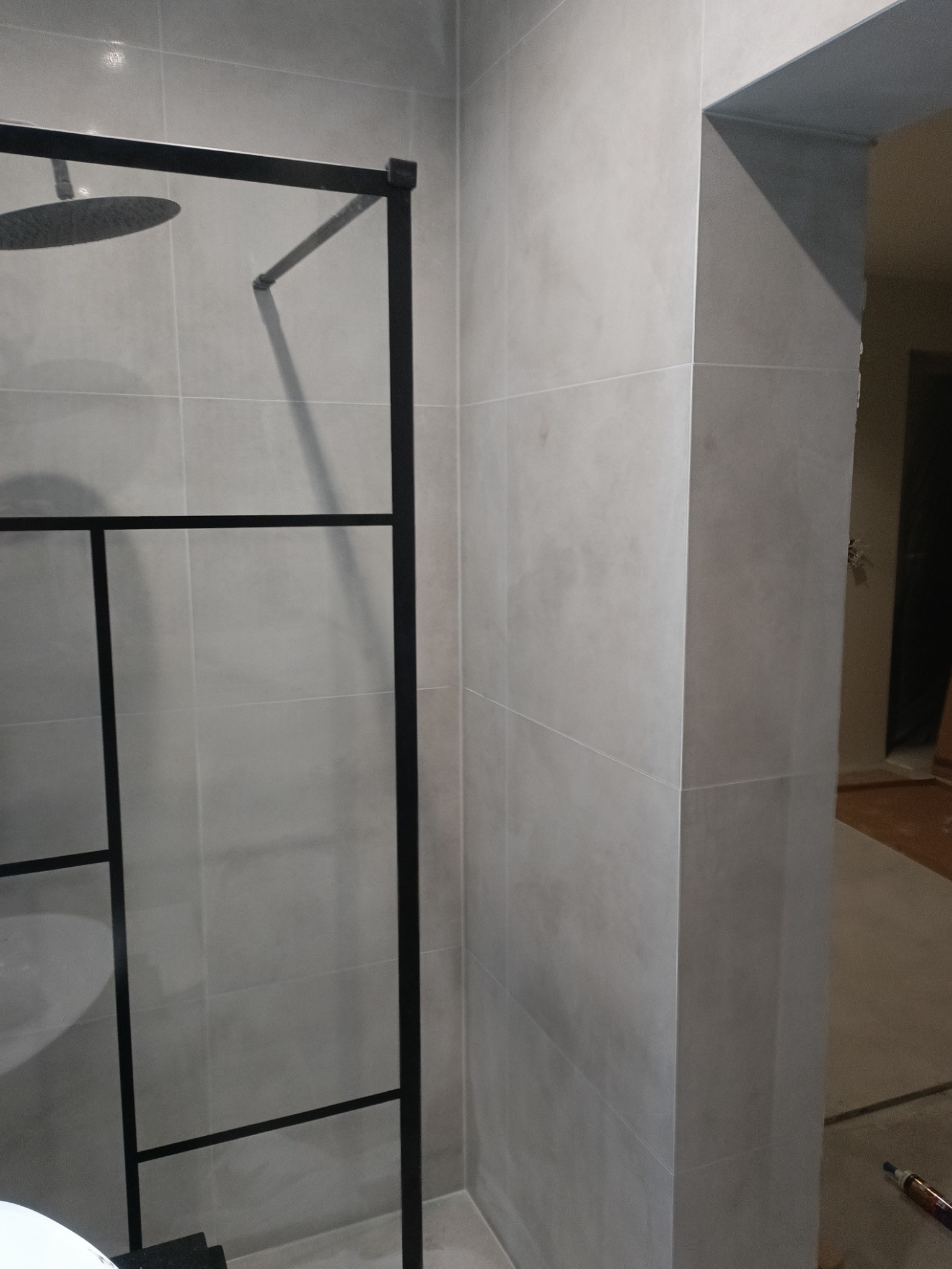 Kabina prysznicowa typu walk-in z czarną ramą i szarymi płytkami na ścianach, widok na fragment łazienki w trakcie wykańczania.
