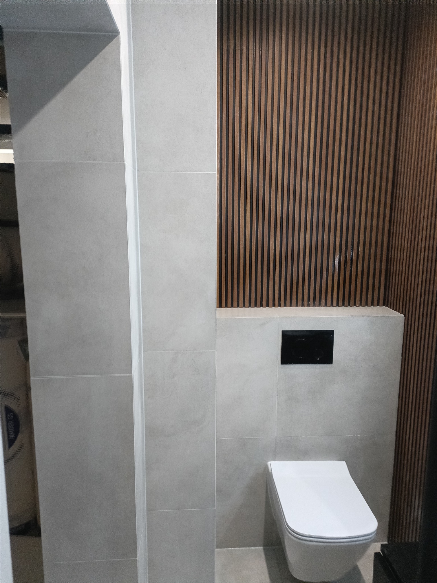 Szare płytki wielkoformatowe na ścianie z ukrytym systemem spłukiwania toalety i dekoracyjnymi drewnianymi lamelami.