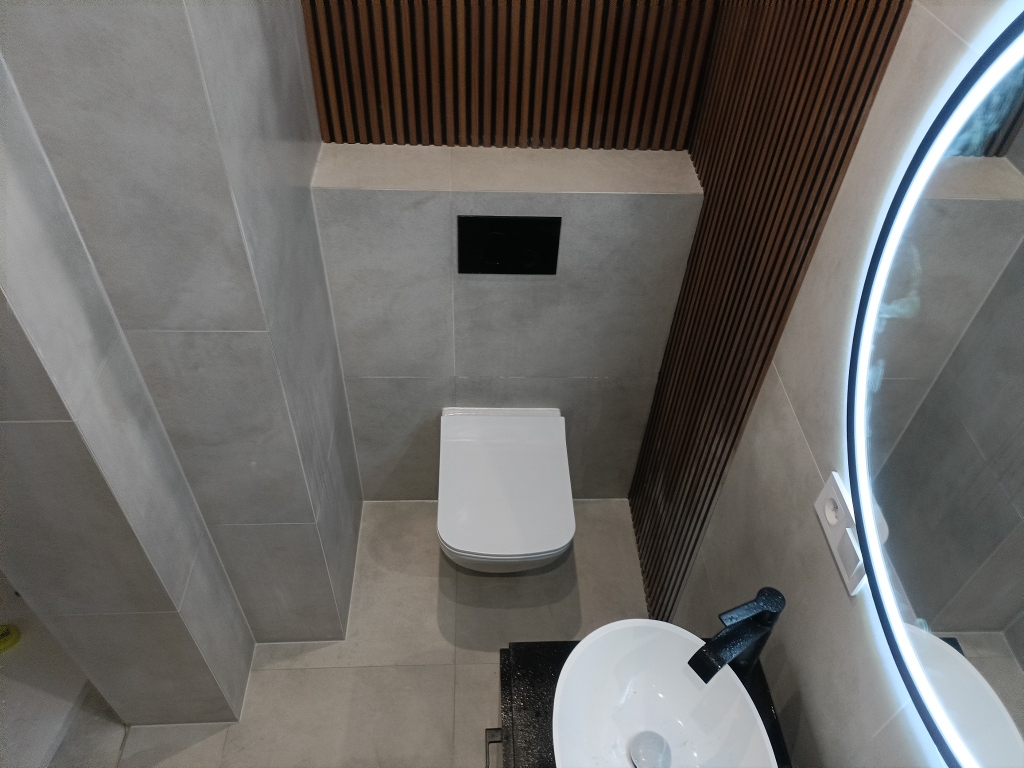 Nowoczesna łazienka z szarymi płytkami, podwieszaną toaletą, umywalką nablatową z czarną armaturą i dekoracyjnymi drewnianymi panelami na ścianie.