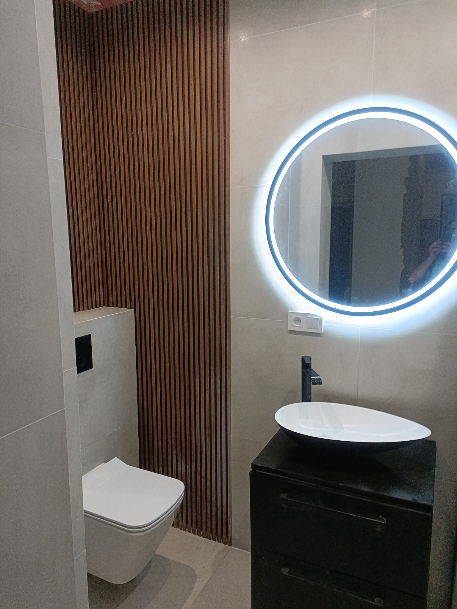 Nowoczesna łazienka z podświetlanym okrągłym lustrem, umywalką nablatową na czarnej szafce, wiszącą toaletą i dekoracyjną ścianką z drewnianych listewek.