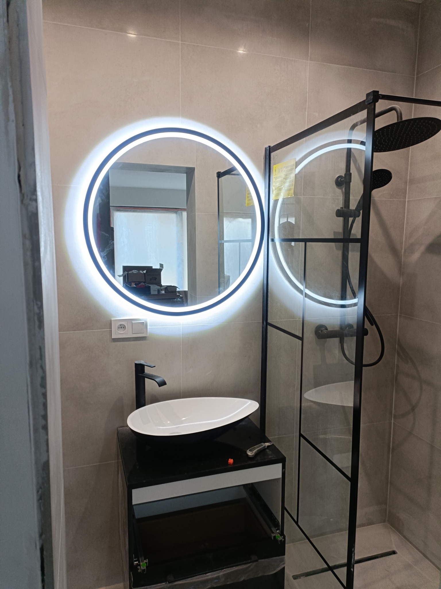 Nowoczesna łazienka z okrągłym lustrem LED, czarną armaturą i kabiną prysznicową typu walk-in z czarnymi profilami; widoczne płytki w odcieniach szarości, minimalistyczny design.
