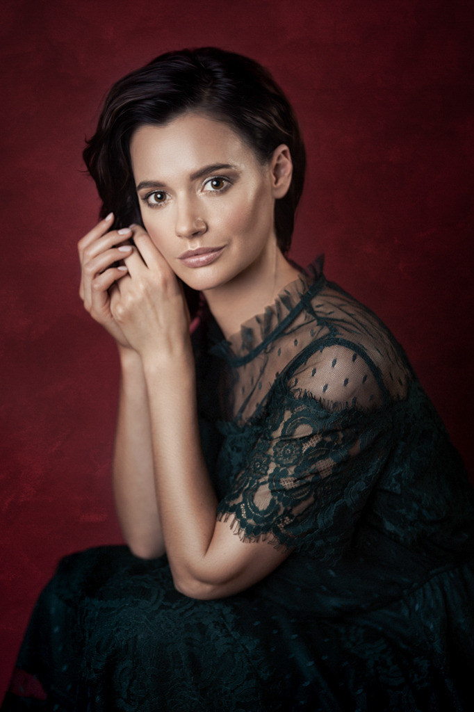 Portret kobiety w zielonej koronkowej sukience na tle czerwonej ściany, dłonie delikatnie dotykają twarzy, subtelny makijaż.