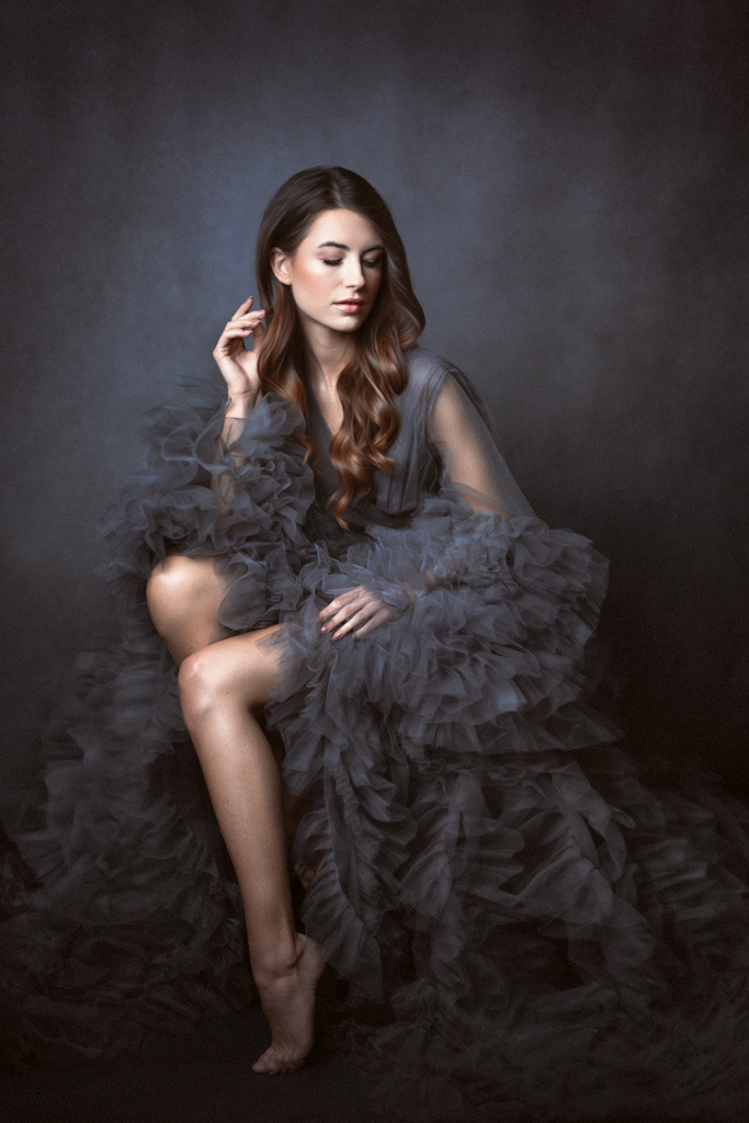 Portret kobiety w zwiewnej, szarej sukni z falbanami, siedzącej na tle ciemnego, teksturowanego tła, z delikatnym oświetleniem podkreślającym jej rysy i fakturę materiału.