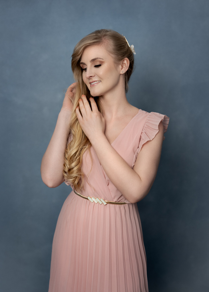Portret młodej kobiety w pudrowo-różowej sukience z plisowanym dołem, delikatnie dotykającej swoich blond loków na tle szaro-niebieskiego płótna.