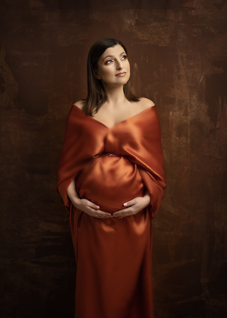 Portret kobiety w ciąży, ubrana w miedzianą, satynową suknię, z rękami na brzuchu, na tle abstrakcyjnego, brązowego tła.
