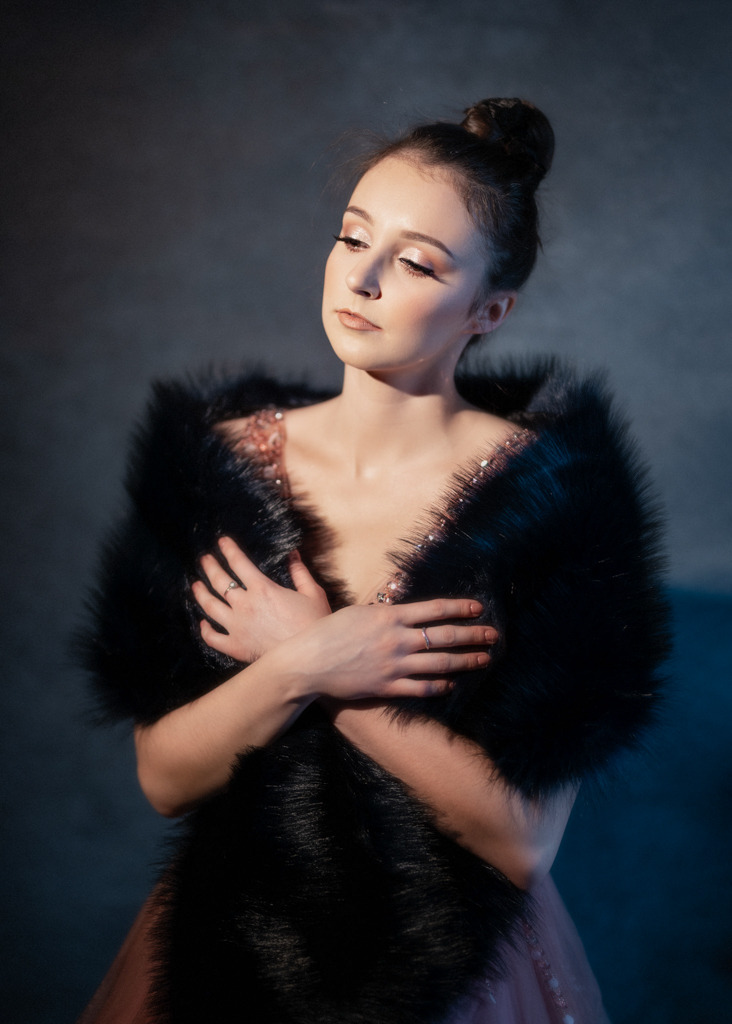 Portret młodej kobiety w eleganckiej sukni z cekinami i czarnym futrzanym szalem, ręce skrzyżowane na piersiach, delikatny makijaż, włosy upięte w kok, patrząca w dół.