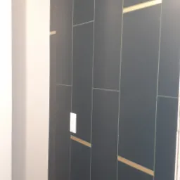 Ciemne, pionowo ułożone płytki w łazience z złotymi akcentami, widoczne wystające przewody i zawory wodne, fragment drewnopodobnej podłogi.