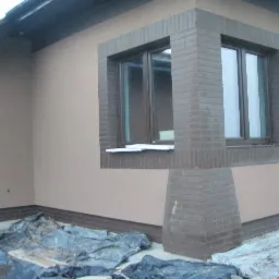 Elewacja domu jednorodzinnego z oknem obramowanym cegłą, widoczne zabezpieczenia folią malarską na ziemi.