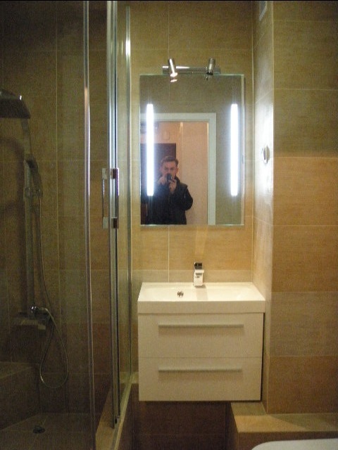 Wykończona łazienka z kabiną prysznicową, umywalką z szafką i lustrem z oświetleniem; odbicie fotografa widoczne w lustrze.
