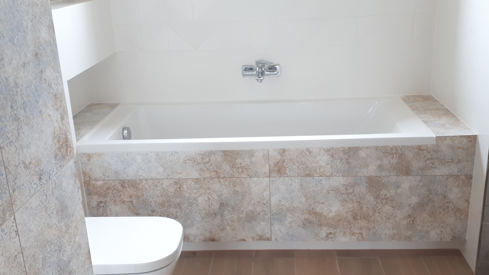 Wykończona łazienka: biała wanna obudowana płytkami w odcieniach beżu i błękitu, biała ceramika sanitarna, jasne płytki ścienne z delikatnym wzorem.