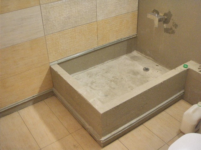 Niedokończony brodzik w łazience z surowego betonu, widoczne płytki na ścianach w odcieniach beżu i brązu, oraz fragment podłogi z płytek ceramicznych, odpływ liniowy w brodziku.